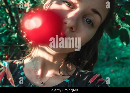 Gros plan portrait d'une jeune femme cueillant des cerises rouges mûres d'un arbre dans un verger en été. Récolte, jardinage, agriculture. Rayons du soleil sur un visage. Banque D'Images