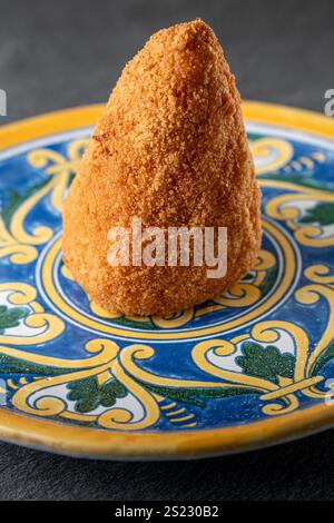 Un seul arancino sicilien ou arancina, une boule de riz traditionnelle avec un revêtement croustillant doré, servi sur une assiette décorative colorée typique de la Sicile Banque D'Images