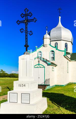Une croix blanche portant le numéro 1934 se dresse devant une église. L'église est blanche et verte, et a un grand clocher. Le ciel est bleu et clair Banque D'Images