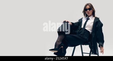 Ambiance confiante et détendue. Femme élégante en costume noir, chemise blanche, cravate, lunettes de soleil et bottes, assise sur la chaise et pose sur fond de studio Banque D'Images