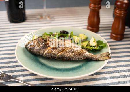 Poisson dorado entier grillé servi avec des herbes fraîches et des légumes dans l'assiette au restaurant. Fumé, tendre et savoureux, ce plat offre un parfait Banque D'Images