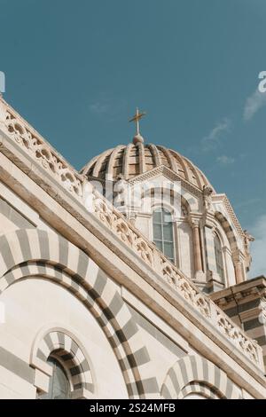 Gros plan de notre-Dame de la Garde à Marseille, France, présentant ses motifs de pierre rayés, son dôme orné et sa croix dorée sur un ciel bleu clair Banque D'Images