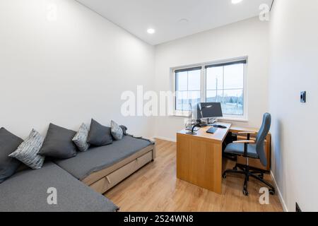 Une configuration de bureau à domicile propre et moderne avec un bureau élégant, une chaise ergonomique et un canapé confortable avec des oreillers, complété par une vue à travers un grand wi Banque D'Images