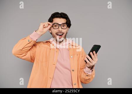 Un jeune homme beau aux cheveux bruns montre de la joie et de la surprise en utilisant son smartphone. Banque D'Images