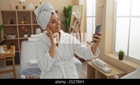 Femme prenant selfie dans le spa portant peignoir et serviette sur la tête dans le centre de bien-être avec éclairage chaud et fournitures de soins de beauté autour Banque D'Images