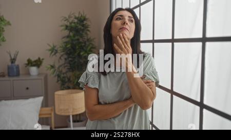 Femme debout dans la chambre portant des vêtements décontractés regardant réfléchi regardant par la grande fenêtre soulignant la beauté et les émotions dans une maison confortable avec natur Banque D'Images