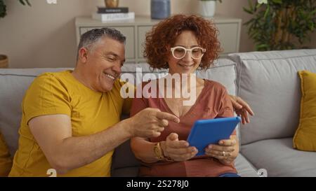 Couple d'âge moyen profitant du temps ensemble dans leur salon, assis sur le canapé et interagissant avec une tablette. Banque D'Images