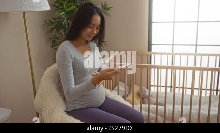 Femme enceinte assise sur une chaise dans une chambre confortable, regardant son téléphone à côté d'un berceau de bébé, illustrant une future mère dans un calme et une domeste Banque D'Images