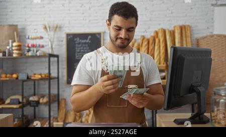 Jeune homme hispanique dans une boulangerie comptant de l'argent derrière un comptoir tout en souriant, entouré de pain frais et de viennoiseries avec un menu au tableau noir dans le bac Banque D'Images