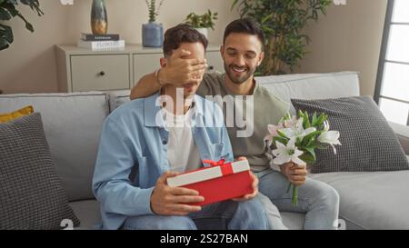 Couple gay assis sur le canapé dans le salon, un homme couvrant les yeux du partenaire avec la main, tenant le cadeau et les fleurs, célébrant l'amour dans la maison confortable interio Banque D'Images