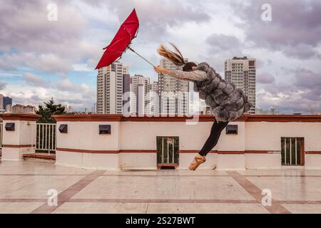 Une ballerine dans un manteau de fourrure saute gracieusement sur un toit, tenant un parapluie rouge contre un paysage urbain venteux avec des immeubles de grande hauteur, Banque D'Images