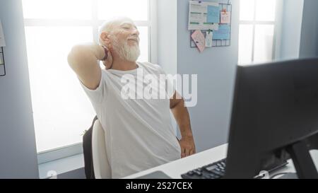 Homme mûr aux cheveux gris dans un bureau intérieur souffrant de douleurs au cou, étirements et griffures alors qu'il était assis à son bureau avec une barbe et un hea chauve Banque D'Images