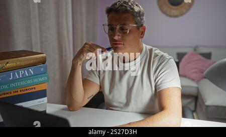 Jeune homme lisant à l'intérieur à la maison tout en réfléchissant à un livre dans le salon avec une pile de livres sur la table. Banque D'Images