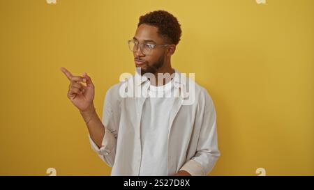 Jeune homme pointant sur le mur de fond jaune isolé portant des lunettes et une chemise décontractée Banque D'Images