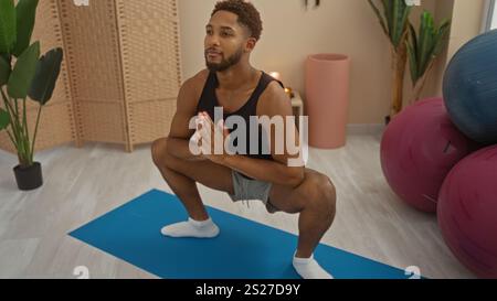 Jeune homme pratiquant le yoga dans un gymnase intérieur, démontrant un squat profond sur un tapis bleu entouré d'équipements de fitness et de plantes Banque D'Images