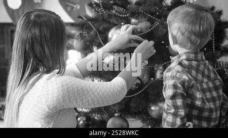 Portrait noir et blanc de tout-petit garçon regardant la mère ornant l'arbre de Noël dans le salon Banque D'Images