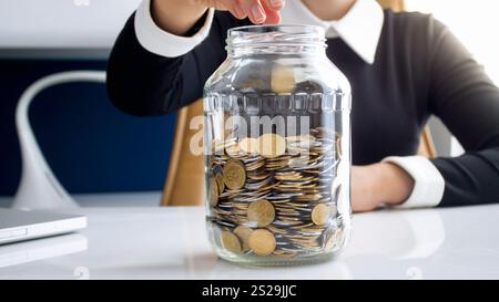 Gros plan photo de jeune femme d'affaires jetant des pièces de monnaie dans un pot en verre avec des économies Banque D'Images