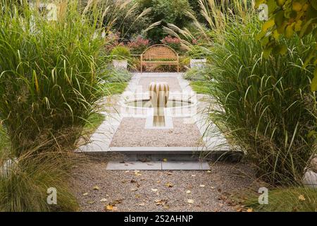 Lit de gravier surélevé avec fontaine d'eau en forme de courge et banc de jardin en treillis en métal brun clair avec Hydrangea paniculata rose «Quick Fire». Banque D'Images