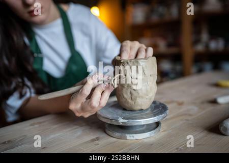 Les mains féminines utilisent habilement l'outil de boucle de poterie pour gratter l'argile et sculpter le motif décoratif sur la tasse Banque D'Images