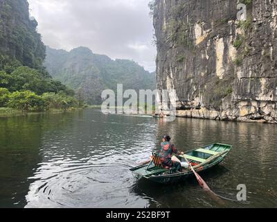 Femme tourne son bateau à aubes à Tam Coc, Ninh Binh, Vietnam. Le bateau pagaie dans une vallée brumeuse, avec des collines spectaculaires de chaque côté. Banque D'Images