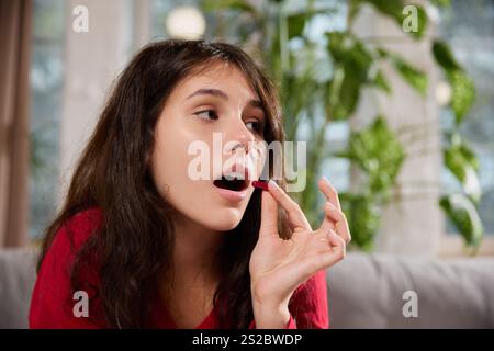 Photo en gros plan d'une jeune femme en pyjama tenant une capsule rouge près de ses lèvres, paraissant concentrée alors qu'elle se prépare à la prendre. Banque D'Images