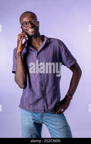Homme africain souriant tout en tenant un smartphone à son oreille dans un studio avec un fond violet pastel Banque D'Images