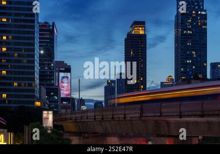 Une prise de vue en exposition longue capture les traînées de lumière dynamiques d'un train BTS qui traverse le paysage urbain de Bangkok à la tombée de la nuit, avec la tour de bus de la ville Banque D'Images
