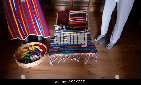 Textiles traditionnels faits à la main avec des motifs rayés colorés affichés aux côtés de paniers tissés et de mannequins sur un plancher en bois dans un intérieur rustique. Banque D'Images