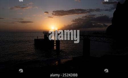 Une silhouette spectaculaire d'une jetée et des figures contre un coucher de soleil incandescent sur l'océan, avec des falaises rocheuses sur le côté. Une vue sereine et pittoresque sur la côte. Banque D'Images