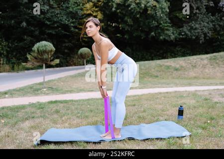 Instructeur de fitness effectuant des exercices d'étirement à l'aide d'une bande de résistance sur un tapis de yoga dans un parc Banque D'Images