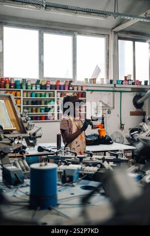 Ouvrière d'imprimerie multiculturelle mélangeant la peinture dans un seau tout en se tenant debout à l'usine entourée de peinture et de sérigraphie presse. Inter Banque D'Images