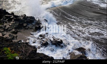 Les vagues s'écrasent contre un rivage rocheux, créant de l'écume marine blanche et des textures d'eau dynamiques, avec un soupçon de verdure sur la côte accidentée. Banque D'Images
