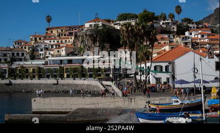 Une charmante ville côtière avec des maisons colorées sur une colline, des palmiers, et un port animé avec des bateaux de pêche et des touristes sous un bleu vif s. Banque D'Images
