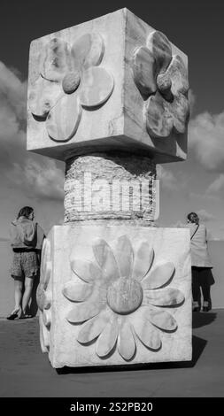 Photo en noir et blanc d'une grande colonne sculpturale présentant des sculptures florales, avec deux personnes debout à proximité contre un ciel nuageux Banque D'Images