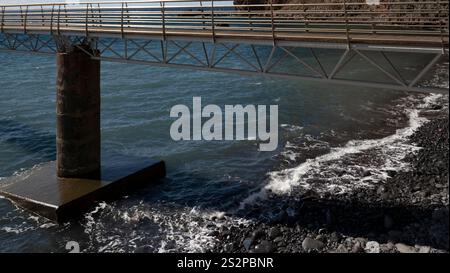 Un pont piétonnier métallique s'étendant sur un rivage rocheux, avec de douces vagues lavant contre les pierres et la mer bleue sous la lumière du soleil. Banque D'Images