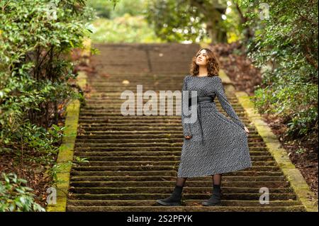 Femme portant une robe florale debout avec confiance sur des escaliers en pierre moussue dans un cadre de forêt verdoyante. Banque D'Images