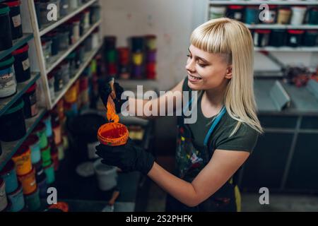 Femme souriante technicien graphique expérimenté debout à l'atelier d'impression avec la boîte d'encre dans les mains, en le mélangeant avec la spatule et en se préparant pour sérigraphie PRI Banque D'Images