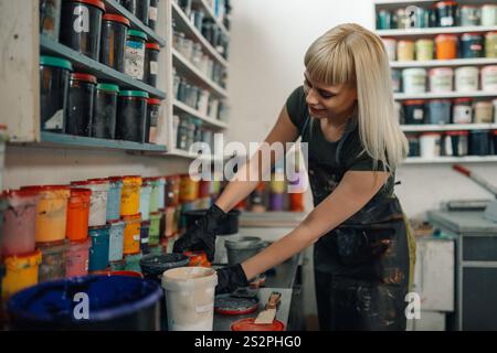 Technicien graphique féminin expérimenté souriant debout à l'atelier d'impression près de l'étagère avec des seaux de peinture, ouvrant et choisissant l'encre pour l'impression sérigraphique Banque D'Images