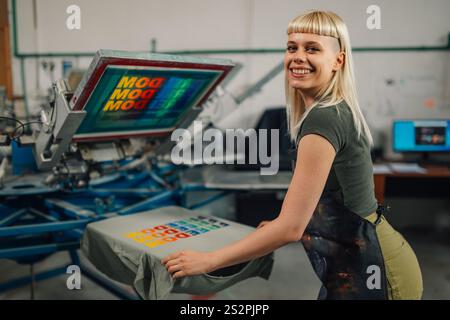 Portrait d'une femme technicien graphique professionnelle sérigraphie sur machine d'impression carrousel et souriant à la caméra. Sourire expérimenté p Banque D'Images