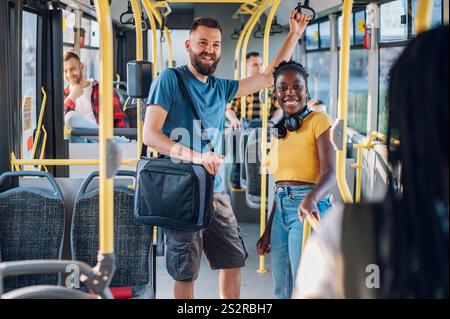 Jeune couple multiracial debout dans un bus en mouvement et allant travailler. Portrait de deux amis souriants divers discutant dans le bus tout en voyageant Banque D'Images