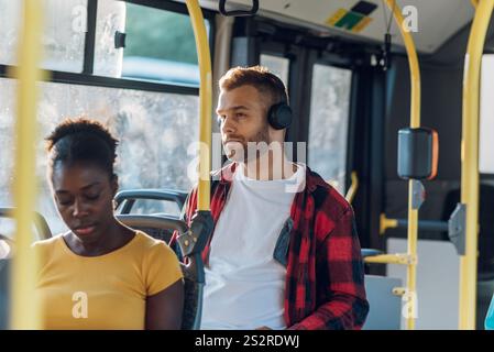 Jeune homme joyeux appréciant la musique pendant son trajet et tenant sur le bar tout en se tenant debout dans un bus. Bel homme prenant le bus pour travailler et utilisant la tête Banque D'Images