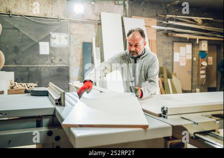 Un ouvrier coupe du bois pour fabriquer des meubles dans son atelier. Un menuisier traite des matériaux et des panneaux en bois sur une machine de découpe Banque D'Images