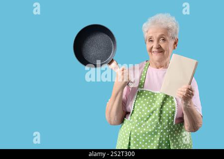 Femme senior dans le tablier avec poêle à frire et livre de recettes sur fond bleu Banque D'Images