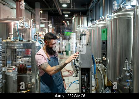 Portrait d'un homme barbu portant un tablier posant dans l'atelier d'une brasserie industrielle moderne. Fabrication de bière avec des récipients métalliques sur le fond. Banque D'Images