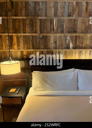 Une chambre confortable et élégante avec un mur d'accent rustique en bois, un lit soigneusement fait avec des draps blancs croustillants, projetant une lueur chaleureuse et accueillante. Minimaliste Banque D'Images