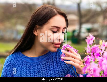 Femme russe dans un pull bleu sentant des fleurs de rhododendron roses dans le Primorsky Krai, Russie. La scène capture l'essence du printemps et du naturel Banque D'Images