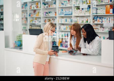 Deux pharmaciennes multiculturelles en blouses de laboratoire debout derrière le comptoir à la pharmacie magasin avec comprimé dans les mains et l'utilisant pour accéder au patient senior Banque D'Images