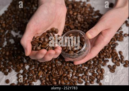 Les mains de la femme choisissent des grains de café torréfiés aromatiques frais dans un petit verre, fond de papier artisanal. Mélange arabica et robusta Banque D'Images