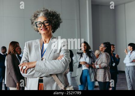Professionnel féminin mature confiant lors d'une conférence d'entreprise, debout et souriant avec divers collègues en arrière-plan engagés dans con Banque D'Images