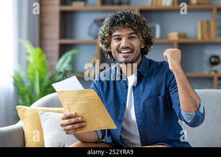 Gagnant de bonnes nouvelles, homme avec l'enveloppe de courrier regardant joyeusement la caméra, assis dans le salon à la maison, a reçu un message de notification par courrier sur de bons résultats. Banque D'Images